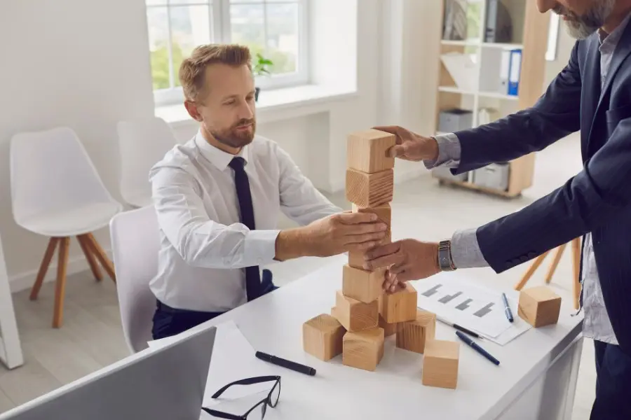 Two professionals in an office building a tower out of wooden blocks on a desk, symbolising teamwork and strategy.