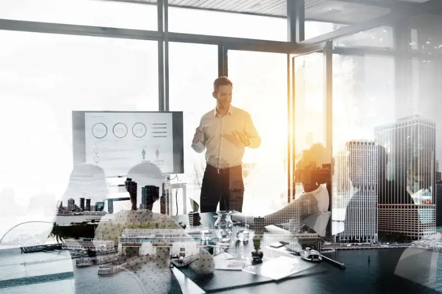 A man presents data on a screen to colleagues seated in a modern glass-walled conference room, with a double-exposure effect blending the meeting with a city skyline.